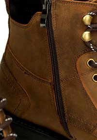 Bota de cuero marrón con cremallera, ojales de metal y cordones marrones. Presenta detalles de costura y una superficie texturizada.