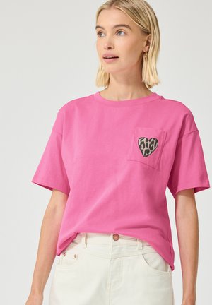 Frau mit kurzen blonden Haaren, die ein pinkes T-Shirt mit einem Leopardenherz auf der Brusttasche und weiße Hosen trägt.