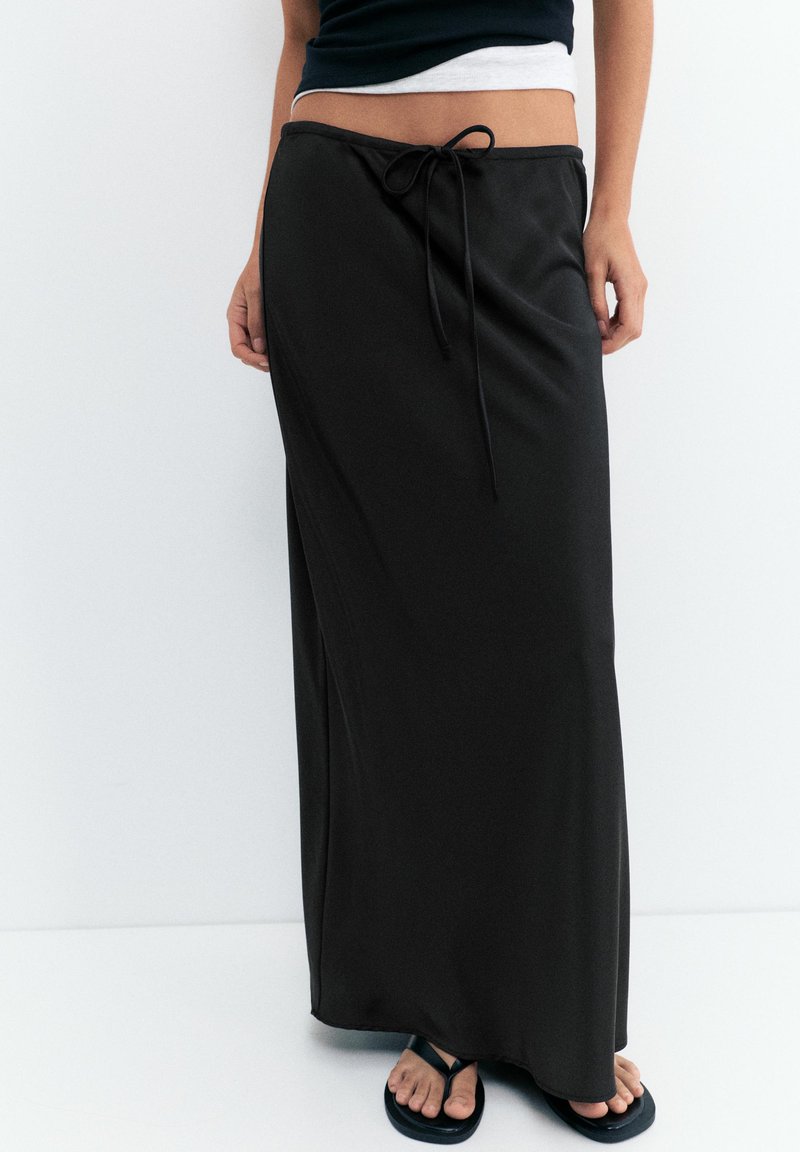 Femme portant une longue jupe noire avec une ceinture nouée, des tongs noires et un haut court noir, debout devant un fond blanc.