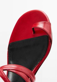 Sandale rouge avec un dessus en cuir lisse et une semelle en cuir noir, comportant deux sangles courbées sur le dessus. Semelle plate, design minimaliste.