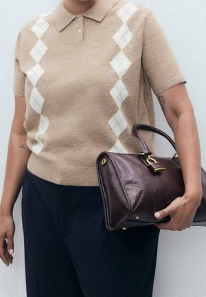 Persona con un suéter beige de manga corta con patrón argyle y pantalones oscuros, sosteniendo un bolso de cuero marrón oscuro con herrajes dorados.