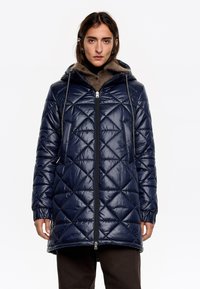 Námornícky prešívaný pufferský kabát s kapucňou, predným zipsom a šnúrkami. Má diamantové vzory a rebrované manžety pre zvýšenie tepla.
