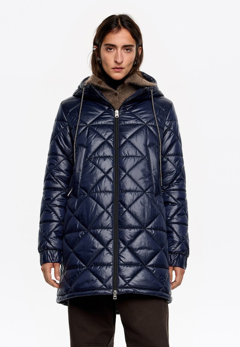 Námornícky prešívaný pufferský kabát s kapucňou, predným zipsom a šnúrkami. Má diamantové vzory a rebrované manžety pre zvýšenie tepla.