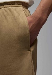 Pantaloni della tuta beige in tessuto testurizzato, con tasca laterale e vita elasticizzata. Dettaglio ravvicinato del tessuto e della mano in tasca.