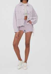 Persona vistiendo una chaqueta de mezclilla oversized color lila claro y shorts deshilachados a juego, top corto blanco y zapatillas blancas frente a un fondo liso.