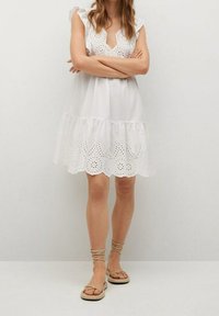 Weißes Baumwollkleid mit V-Ausschnitt, gerafften Kapussärmeln und gewelltem Spitzenbordüre am Saum. Kombiniert mit beigen flachen Sandalen.