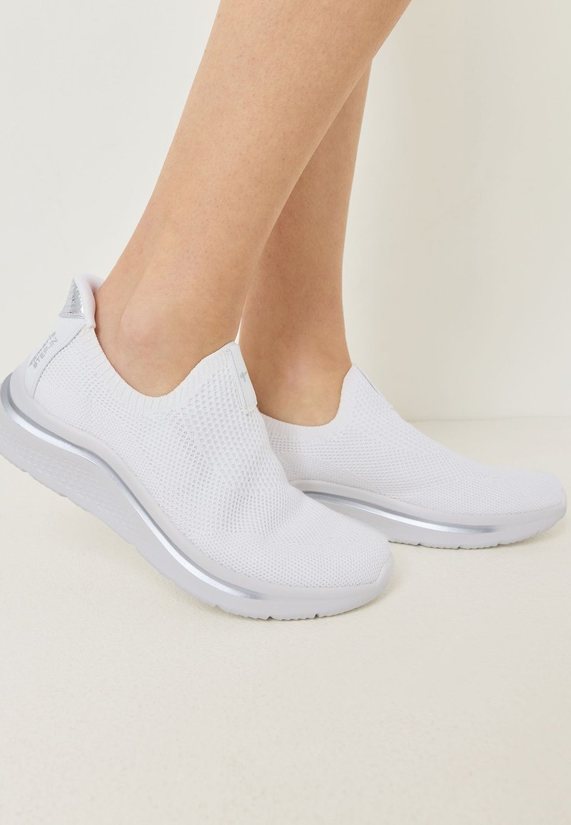 Jambes féminines portant des baskets en maille blanches sans lacets, avec des semelles épaisses et des accents argentés sur un fond neutre.
