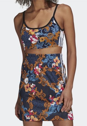 Kobieta ubrana w dopasowany sportowy biustonosz i spódnicę z wzorami kwiatowymi w kolorach niebieskim, pomarańczowym, czerwonym i białym, oba elementy z logo Adidas i trzema białymi paskami.