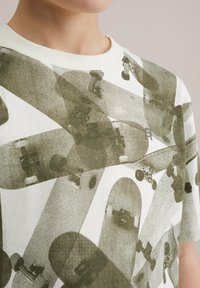Camiseta de algodón blanca de manga corta con un patrón gráfico verde de patinetas superpuestas y un pequeño logo en el frente.