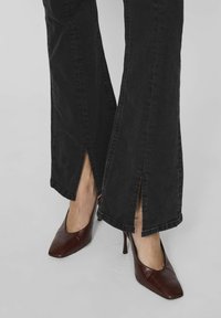 Melni zvēru džinsi ar sānu šķēlumiem pie apakšējiem malām, apvienoti ar brūnām augstpapēžu kurpēm ar vērsto purngalu. Džinsu audums ar gludu tekstūru.