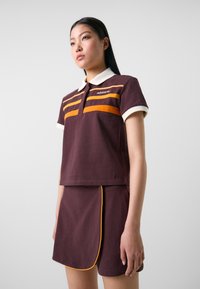 Burgundi pólóing fehér gallérral és narancssárga csíkokkal, párosítva egy illő burgundi szoknyával, amely sárga részletekkel és oldalsó gombdíszítéssel rendelkezik.