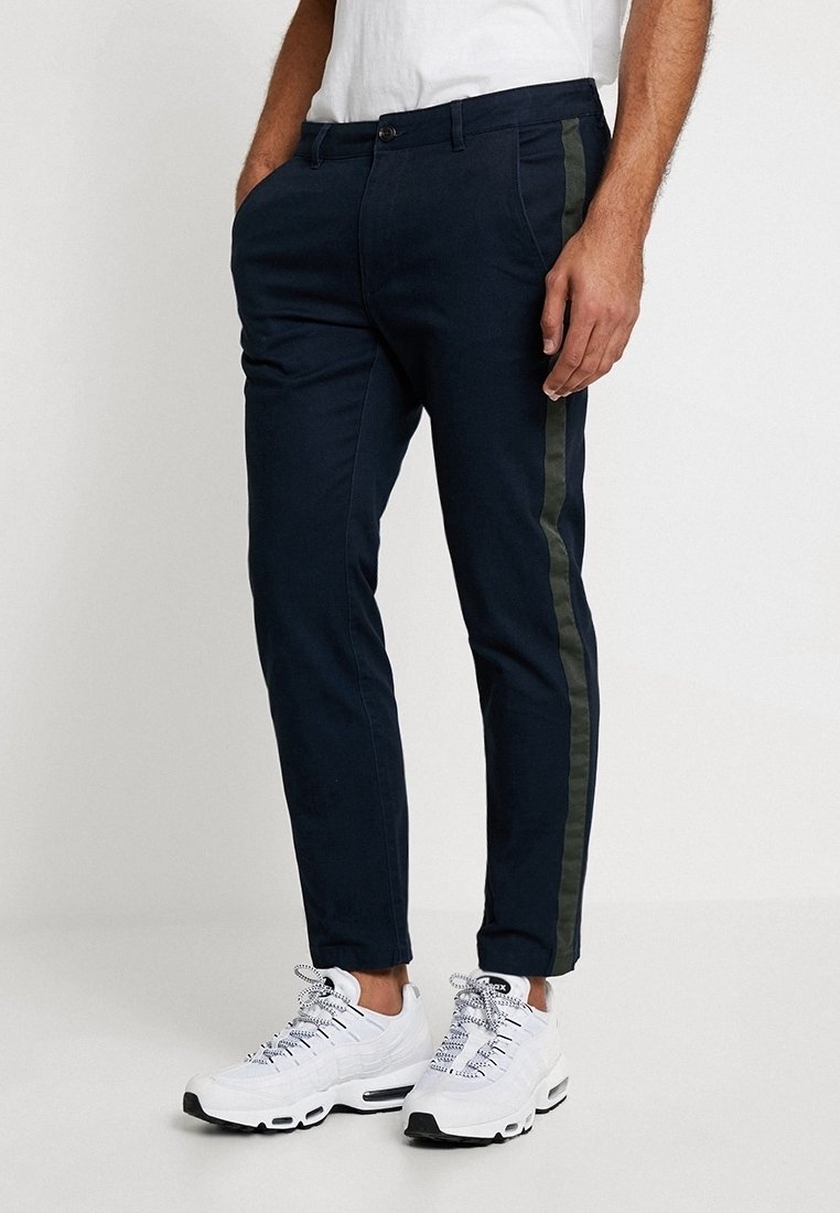 Pantalon extensible bleu marine avec des bandes latérales vert olive, coupe fuselée, passants de ceinture et poches avant. Associé à des baskets blanches.
