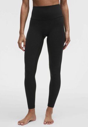 Kvinde iført sorte leggings med høj talje, stående barfodet på en neutral baggrund, armene afslappet langs siderne, beskåret ved midten af overkroppen.