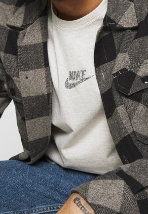 Graues und schwarzes kariertes Flanellhemd über einem hellgrauen T-Shirt mit Nike-Logo; deutliche Textur im Stoff; sichtbare Jeans.