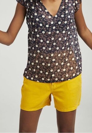 Kobieta ubrana w ciemną, kwiecistą bluzkę z krótkim rękawem w białe kwiaty i jasne żółte szorty, stojąca z lekko podniesionymi ramionami.