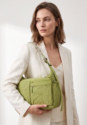 Vrouw in een beige pak houdt een lichtgroene GMY-tas met een ritssluiting en een knoop aan de schouderriem, kijkt naar de zijkant.