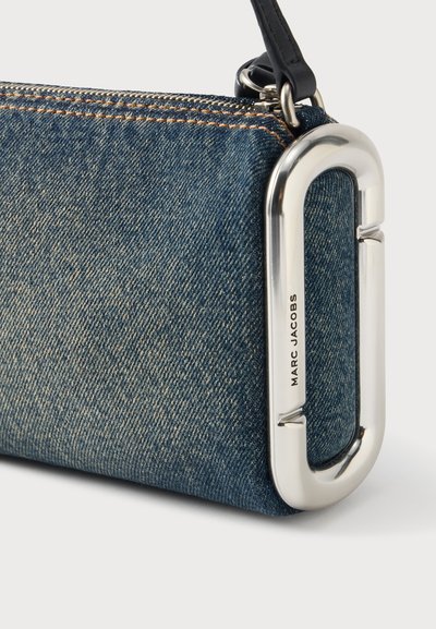Sac bracelet en denim avec coutures orange, fermeture éclair en métal et mousqueton ovale argenté marqué Marc Jacobs sur fond blanc.