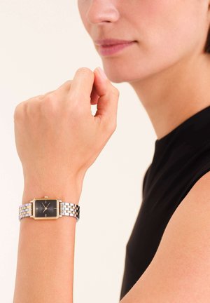 Montre carrée avec un cadran noir, des accents dorés, et un bracelet en métal argenté et doré. Portée sur un poignet contre un fond neutre.