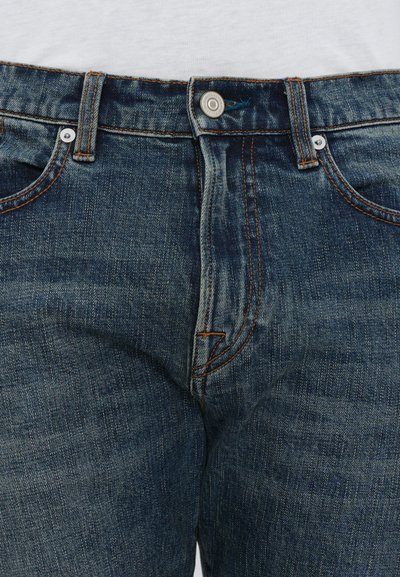 Jean en denim bleu foncé avec fermeture à bouton, présentant des coutures orange, cinq poches et une texture légèrement décolorée.