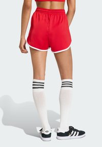 Crvene sportske kratke hlače s bijelim obrubom, elastičnim pojasom i opuštenim krojem, uparene s bijelim čarapama do koljena s crnim prugama i crnim tenisicama.