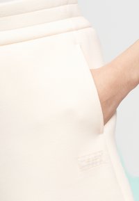 Joggers couleur pêche claire en tissu doux, avec une poche latérale et une petite étiquette sur la ceinture. Texture lisse et coupe décontractée.