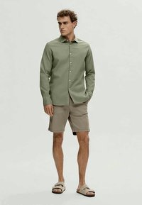 Man wearing light green button-up shirt, beige shorts, and beige sandals, standing with one hand in pocket against plain background.