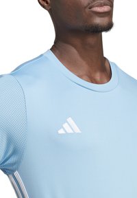 Ljusblå atletisk tröja i fukttransporterande material. Har en rund halsringning, nätpaneler på sidorna och en vit logotyp på bröstet.