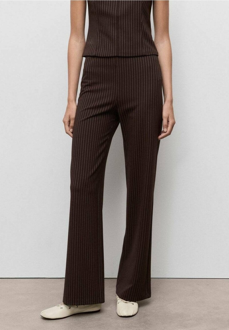 Femme portant un pantalon évasé marron foncé à fines rayures associé à un haut sans manches assorti et des chaussures plates couleur crème, debout contre un mur uni.