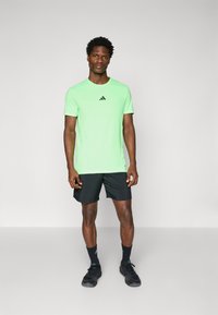 Neon groene sportt-stijl met korte mouwen en zwart Adidas-logo op de borst, gecombineerd met zwarte shorts en zwarte schoenen.