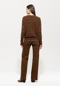 Brauner, gestrickter Pullover mit horizontalen Streifen, kombiniert mit braunen, aus Wildleder gefertigten, weit geschnittenen Hosen. Das Model steht mit dem Rücken zur Kamera.