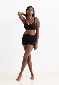 Frau, die barfuß vor einem weißen Hintergrund steht, in einem schwarzen nahtlosen BH und Shorts, mit einem Arm hinter ihrem Kopf erhoben.
