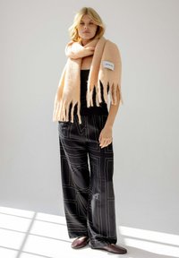 Écharpe rose à franges en tissu doux, portée sur un haut noir. Pantalon noir avec des lignes géométriques blanches. Design simple et minimaliste.