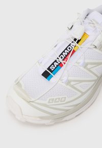 Zapatilla deportiva blanca con parte superior de malla, acentos reflectantes, branding en negro y colorido, y un diseño único de suela para tracción.