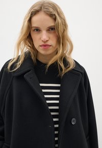 Black wool coat with a wide collar, single button closure, and oversized fit, layered over a navy and white striped sweater.