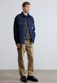 Μπουφάν τζιν σε σκούρο μπλε με δύο τσέπες στο στήθος, φορεμένο πάνω από γκρι μπλούζα φούτερ, σετ με μπεζ παντελόνια και μαύρα παπούτσια. Μινιμαλιστικός σχεδιασμός.