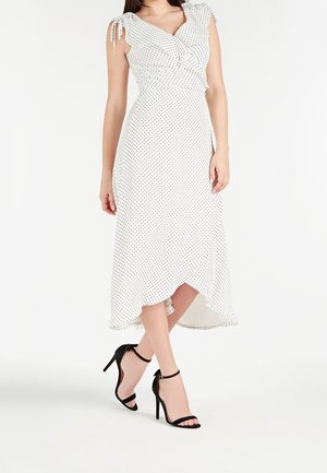 Weißes Midikleid mit schwarzen Punkten, mit Rüschenärmeln, die an den Schultern gebunden sind, und asymmetrischem Saum, kombiniert mit schwarzen High-Heel-Sandaletten mit Knöchelriemen.