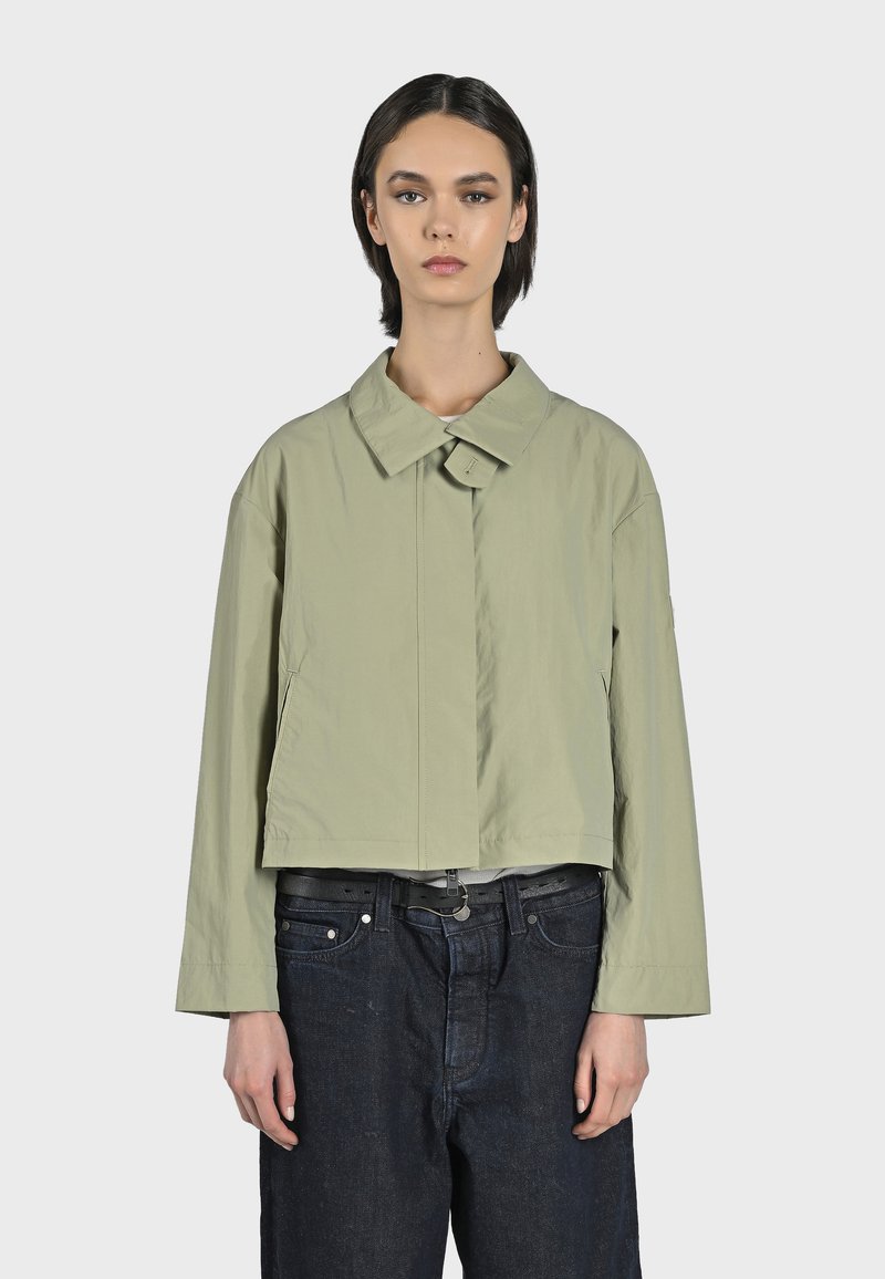 Kort, ljusgrön jacka med en stor krage, framknäppning och långa ärmar. Bärs över mörka jeans med ett svart bälte. Slät textur.