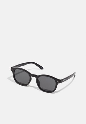Óculos de sol pretos em formato oval com lentes escuras, armações grossas e detalhes em ponto prateado nas laterais. Material plástico suave, design contemporâneo.