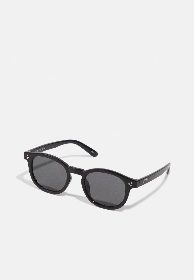 Schwarze ovale Sonnenbrillen mit dunklen Gläsern, dicken Rahmen und silbernen Punktekzenten an den Seiten. Glattes Plastikmaterial, zeitgemäßes Design.