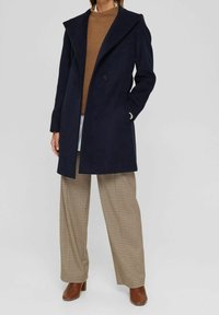 Navyblauw wollen jas met een brede piekkragen, twee knopen en zijzakken, gecombineerd met een bruine gebreide trui en geruite broek.