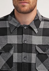 Camisa de franela a cuadros en negro y gris con cuello, dos bolsillos en el pecho y cierres de botones. La tela tiene una textura suave y detalles de costura visibles.