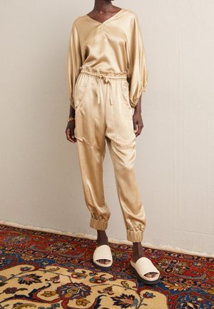 Personne portant un haut en satin doré et un pantalon à cordon assorti avec des poignets élastiques, des sandales beiges à enfiler, debout sur un tapis à motifs.