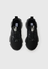 Une paire de baskets noires en mesh et cuir verni avec des lacets élastiques sans nouage et des poignées de marque sur un fond blanc.