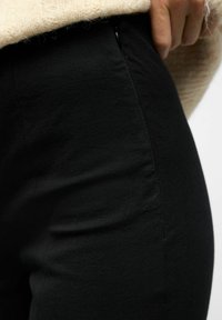 Detailní záběr osoby v černých kalhotách a krémovém svetru, s jednou rukou opřenou u pasu.