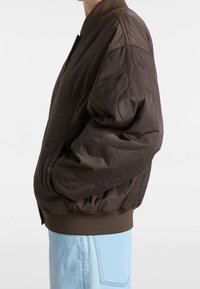 Brązowa pikowana kurtka bomberka z prążkowanym kołnierzem i mankietami, posiadająca boczne kieszenie i luźny fason. Materiał jest gładki i błyszczący.