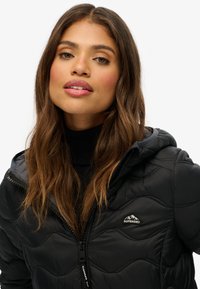 Vrouw met lang bruin haar draagt een zwart gewatteerd Superdry-jack met capuchon over een zwart topje, kijkt iets omhoog en vooruit tegen een effen achtergrond.