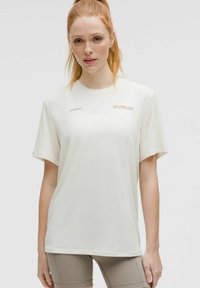 Eine rothaarige Frau trägt ein lockeres weißes Lululemon-T-Shirt und taupefarbene Sportshorts und steht vor einem schlichten hellen Hintergrund.