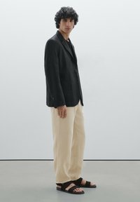 Blazer en lin noir à deux boutons, associé à un pantalon large beige et des sandales à brides noires. Texture lisse et design ajusté.