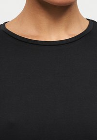 Κοντινό πλάνο στον λαιμό και το ανώτερο μέρος του στήθους ενός ατόμου που φοράει απλό μαύρο μπλουζάκι με στρογγυλή λαιμόκοψη σε ανοιχτό φόντο.