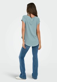 Frau von hinten zu sehen, die ein hellblaues Kurzarmoberteil mit einem Reißverschluss auf dem Rücken, blaue Schlagjeans und weiße Sneakers trägt, vor einem einfarbigen Hintergrund.